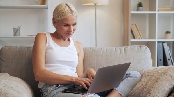 Internet leisure. Home online. Weekend gadget. Relaxed mature woman using laptop on cozy couch at modern interior with free space. - Фото, изображение