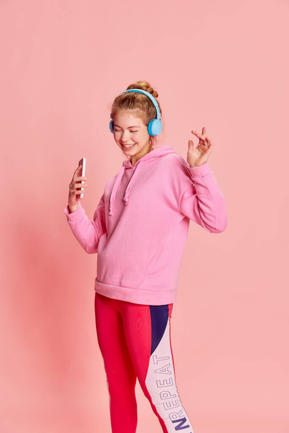Portré a táncoló fiatal lányról, aki fülhallgatót visel, és rózsaszín háttérrel rendelkező zenét hallgat. Koncepciója zene, sport, emberi érzelmek, diák élet, divat, szabadidő - Fotó, kép