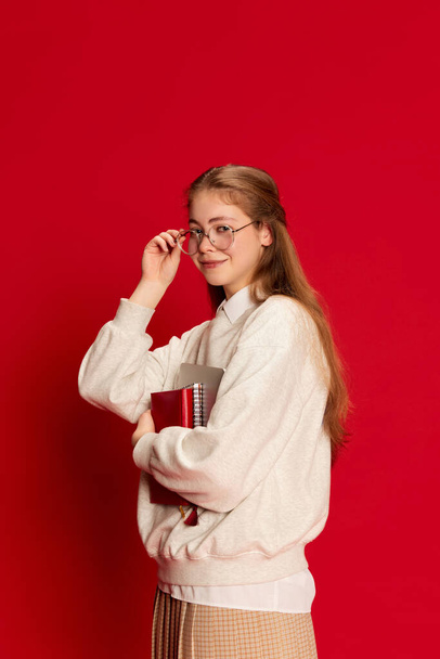 Portré fiatal diáklányról, aki egyenruhát visel, jegyzetfüzetekkel a kezében, és vörös háttér felett nézi a kamerát. Fogalom a hallgatói élet, tanulás, iskola, külföldi oktatás, tanulás - Fotó, kép