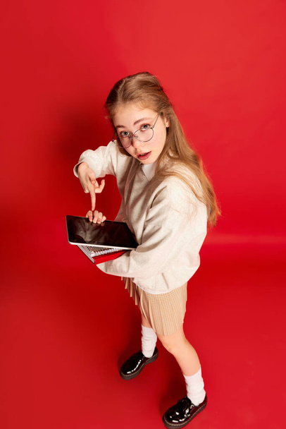 Photo of pupil girl wearing discreet suit , holding notebooks, and pointing with finger on empty tablet screen over red background. Concept of student life, studying, learning, abroad educational - Photo, image