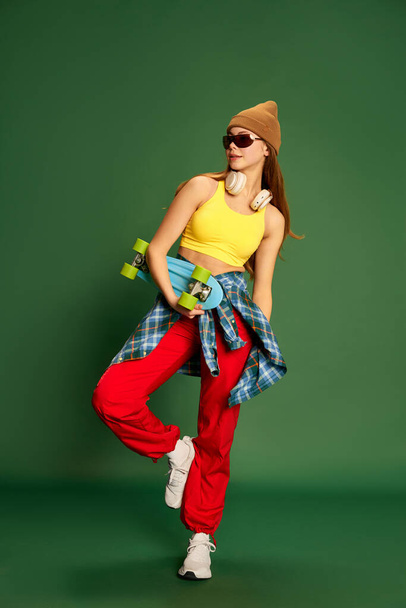 Full-length photo of skateboarding girl wearing stylish clothes and holding skateboard in hands over green background. Concept of youth, fashion, sport, hooby, fun, activity and leisure time - Photo, image