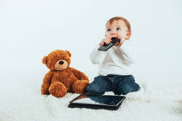 Foto de estúdio com o fundo branco do rosto de um bebê com um celular na boca ao lado de um ursinho de pelúcia e um tablet
. - Foto, Imagem