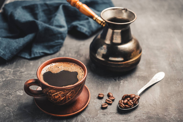 Bebida de café preta em uma xícara de barro, panela de café jezve turca e grãos de café em uma colher no fundo escuro. - Foto, Imagem