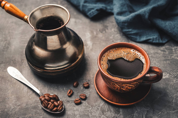 Černý kávový nápoj v hliněném šálku, turecký kávový hrnec a kávová zrna v lžíci na tmavém pozadí. - Fotografie, Obrázek
