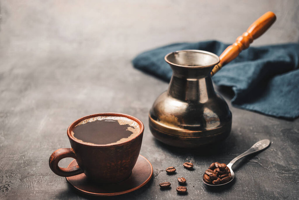 Bebida de café negro en una taza de arcilla, jezve turco cafetera y granos de café en una cuchara sobre fondo oscuro. - Foto, Imagen