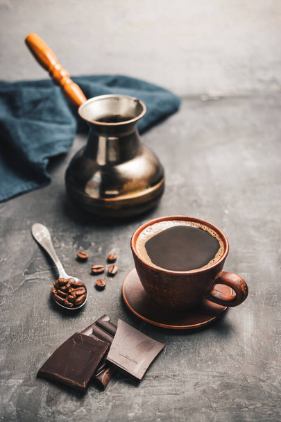 Boisson au café noir dans une tasse d'argile, cafetière turque jezve, morceaux de chocolat et grains de café dans une cuillère sur fond sombre. - Photo, image