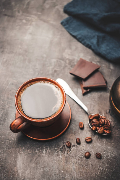 Μαύρο ρόφημα καφέ σε πήλινο κύπελλο, κομμάτια σοκολάτας και κόκκους καφέ σε κουτάλι σε σκούρο φόντο. - Φωτογραφία, εικόνα