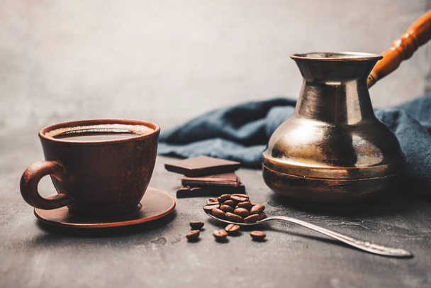 Bebida de café preta em uma xícara de barro, panela de café jezve turca, pedaços de chocolate e grãos de café em uma colher no fundo escuro. - Foto, Imagem