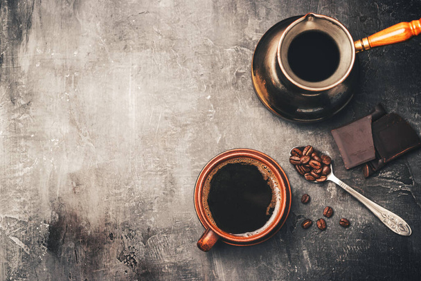 Μαύρο καφέ ποτό σε πήλινο κύπελλο, τούρκικη jezve καφετιέρα, κομμάτια σοκολάτας και κόκκους καφέ σε ένα κουτάλι σε σκούρο φόντο, top view. - Φωτογραφία, εικόνα
