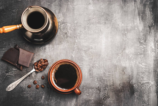 Zwarte koffie drinken in een klei kopje, turkse jezve koffiepot, chocolade stukken en koffiebonen in een lepel op donkere achtergrond, bovenaanzicht. - Foto, afbeelding