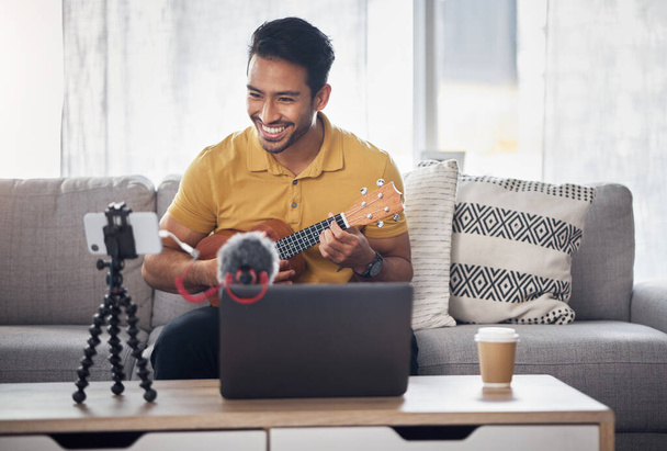 Guitar, streaming and phone with a man talking online to coach during live lesson. Asian male influencer happy on home sofa with a ukulele as content creator teaching music on education podcast blog. - 写真・画像