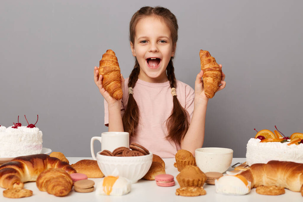 Indoor shot of excited amazed little girl with pigtails sitting at table isolated over gray background, screaming with happiness and excitement with croissant. - Fotó, kép