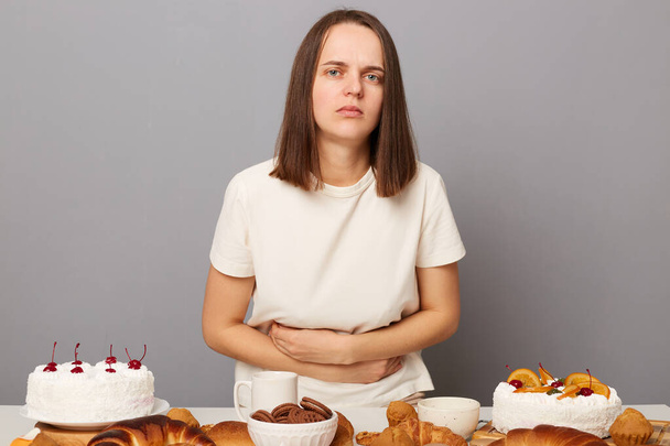 Portrait of sick ill unhealthy woman with brown hair sitting at table isolated over gray background, eating lots sugary homemade bakery, feels stomachache, pain in tummy. - Fotografie, Obrázek