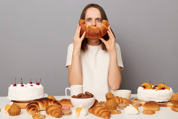 Indoor shot of funny serious woman with brown hair sitting at table isolated over gray background, having dinner, posing among sweet pastry, covering mouth with bagel, having fun. - Zdjęcie, obraz