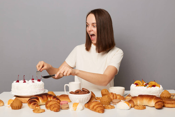 Photo of amazed surprised excited woman with brown hair sitting at table isolated over gray background, holding fork and knife and eating white delicious cake. - Foto, afbeelding