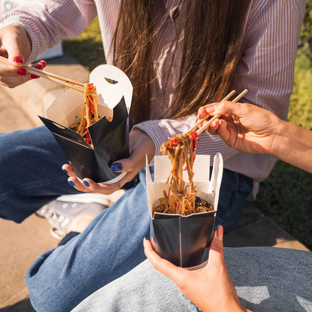 young girls, girlfriends eat asian food with wok noodles, using chopsticks, taking out takeaway food, paper box with takeaway noodles - Photo, image
