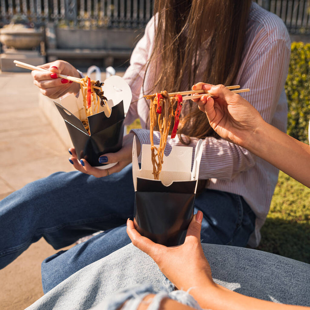 young girls, girlfriends eat asian food with wok noodles, using chopsticks, taking out takeaway food, paper box with takeaway noodles - 写真・画像