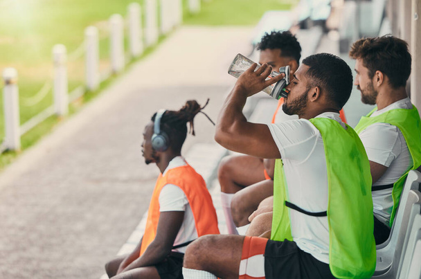 Team, rugby men and fitness on bench with drinking water, relax or sitting with diversity, solidarity or health. Group teamwork, university or professional sports at stadium for game, friends or goal. - Photo, Image