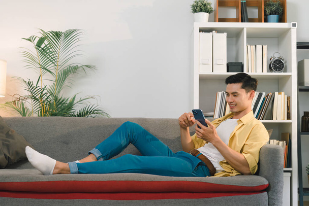 En una acogedora sala de estar en casa, un hombre guapo se sienta en el sofá, absorto en la mensajería en su teléfono inteligente con dedos hábiles - Foto, Imagen