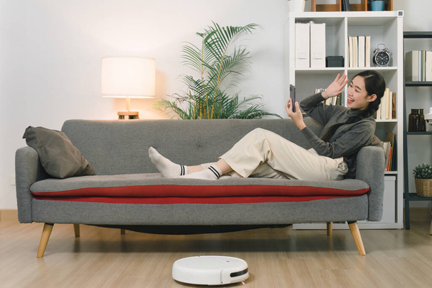 Очаровательная азиатка пользуется смартфоном, расслабляясь дома на диване - Фото, изображение