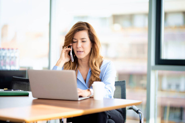 Candid shot of a mid aged professional woman wearing shirt and using laptop while working in a modern office. Attractive businesswoman sitting at desk and having a business call. - Photo, image