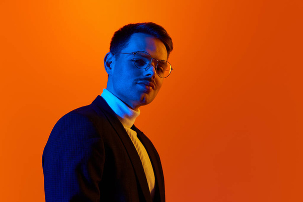 Confident look. Portrait of young man in jacket and glasses posing against orange studio background in neon light. Concept of youth, emotions, facial expression, lifestyle, sales. Ad - Foto, imagen