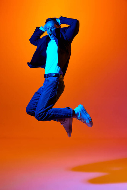 Full-length portrait of young emotive man in stylish casual clothes emotinally jumping against orange studio background in neon light.. Concept of youth, emotions, facial expression, lifestyle. Ad - Foto, imagen