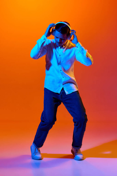 Full-length portrait of young man in white shirt dancing and listening to music in headphones against orange studio background in neon light. Concept of youth, emotions, facial expression, lifestyle - Foto, Imagem