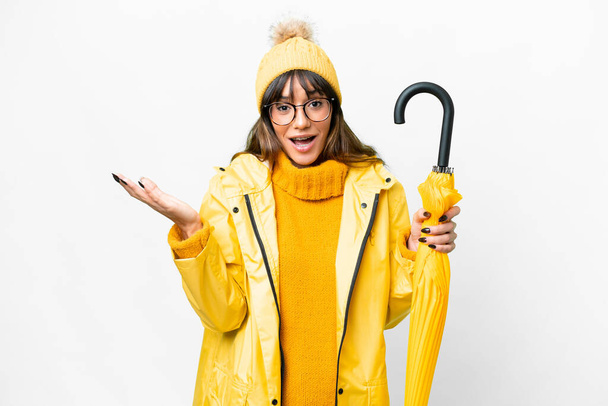 Young caucasian woman with rainproof coat and umbrella over isolated white background with shocked facial expression - Foto, Imagem