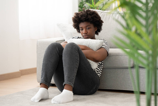 Triste donna afroamericana seduta sul pavimento e che abbraccia cuscino a casa. - Foto, immagini