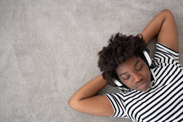 Χαλαρώνοντας Αφρο-Αμερικανός ακούγοντας μουσική στα ακουστικά και ονειρεύεται με χώρο αντίγραφο. Νεαρή μαύρη γυναίκα ξαπλωμένη στο χαλί - Φωτογραφία, εικόνα