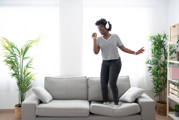 Χαρούμενο αφροαμερικάνικο τραγούδι στο σπίτι. Μαύρη γυναίκα στέκεται στον καναπέ ακούγοντας μουσική - Φωτογραφία, εικόνα