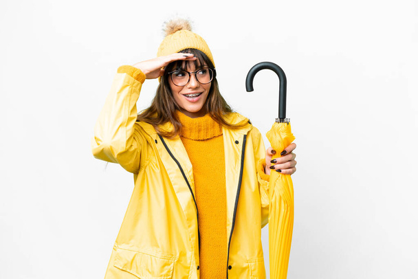 Young caucasian woman with rainproof coat and umbrella over isolated white background doing surprise gesture while looking to the side - Foto, imagen