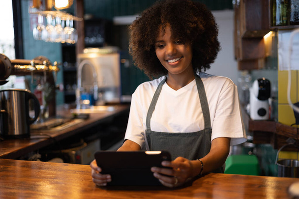 Junge afrikanische Kellnerin mit Afro-Haaren lächelt, während sie Bestellungen auf dem Tablet prüft. - Foto, Bild
