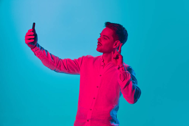 Portrait of young man in shirt taking selfie with mobile phone against blue studio background in pink neon light. Social media. Concept of youth, emotions, facial expression, lifestyle. Ad - Фото, изображение