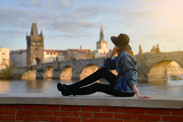 Stylish young beautiful woman earing black hat in Prague with Charles Bridge on background. Elegant retro lady fine art portrait. - Photo, image