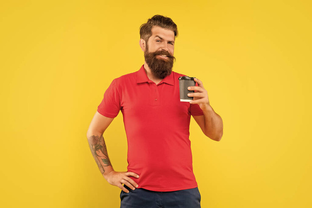 Happy man in casual red tshirt holding disposable cup with arm akimbo yellow background, takeaway coffee. - Фото, изображение