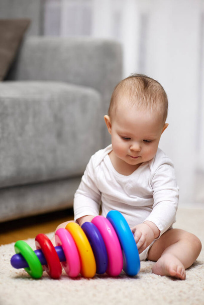 Cute baby girl playing with colorful toy pyramid sitting on carpet at home. Early development for kids. - Foto, imagen