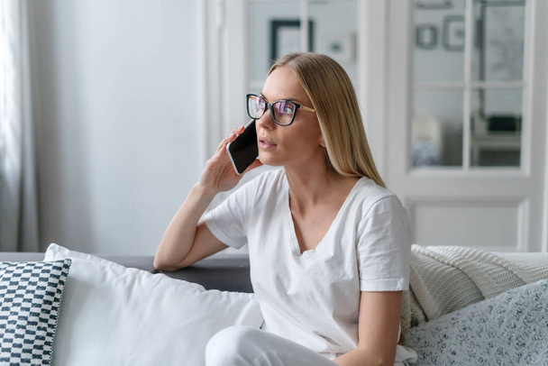 mujer en gafas mantener el teléfono celular en la mano, hablar y tener conversación mientras está sentado en el sofá en la sala de estar, chica discutir problemas de trabajo - Foto, imagen