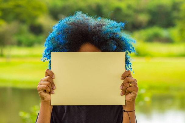 Μια νεαρή γυναίκα, με βαμμένα τα μαλλιά μπλε, το πρόσωπό της κρυμμένο πίσω από μια λευκή αφίσα, με ένα τοπίο στο βάθος. - Φωτογραφία, εικόνα