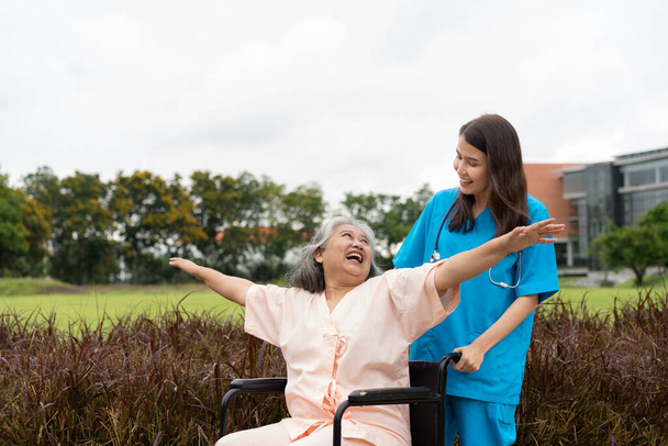 Asian careful caregiver or nurse and the happy patient in a wheelchair are walking in the garden. Concept of happy retirement with care from a caregiver and Savings and senior health insurance. - Foto, afbeelding