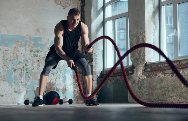 Strength and endurance. Young muscular man doing workout, training with battle ropes indoors. Functional training exercises. Concept of sportive lifestyle, body care, fitness, hobby, health - Foto, Imagem