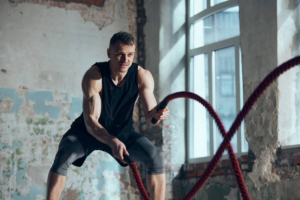 Dynamic image of young sportive man with strong muscular fit body training, doing exercises with battle ropes indoors. Concept of sportive lifestyle, body care, fitness, hobby, health, motion - Foto, Imagem