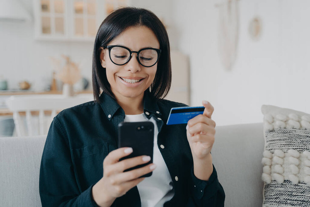 Smiling female holding bank credit card and smartphone. Young woman pays for purchases in online store using banking service, shopping, making secure payment, sitting on sofa at home. E-commerce. - Foto, imagen