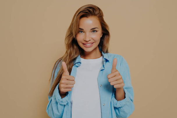 Well done. Happy young female wearing blue long sleeves shirt over white tshirt making thumb up sign with both hands and smiles cheerfully, showing her support and respect - Foto, Bild