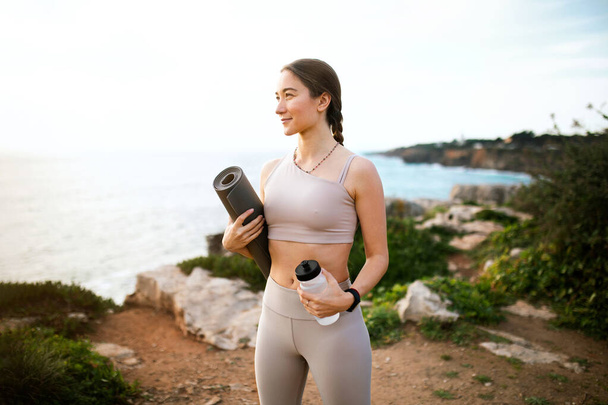 Smiling slim millennial european female in sportswear with mat and bottle of water enjoy training on beach near sea, outdoor. Health care, body care, weight loss and active lifestyle - Photo, Image