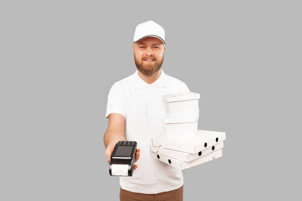 Delivery man wearing white is holding POS terminal and food delivery boxes over grey background. - Photo, Image