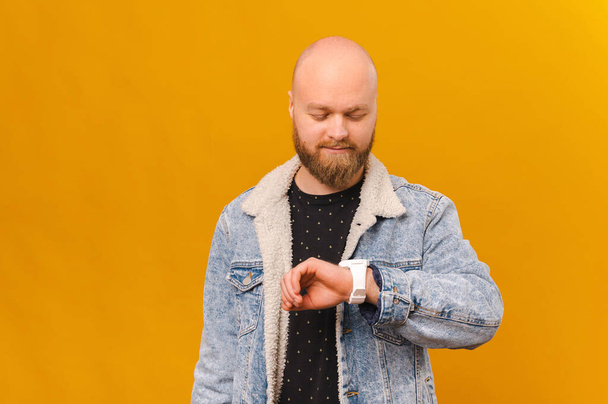 Cool smiling bearded bald man wearing jeans jacket is checking hi watch over yellow background. - Foto, immagini