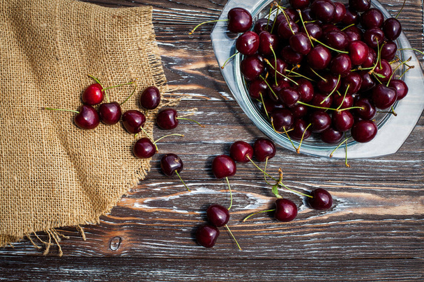 A lot of ripe red cherries on twigs lie in a glass plate on a dark blue wooden table with a striped texture, covered with cloth. - Foto, afbeelding