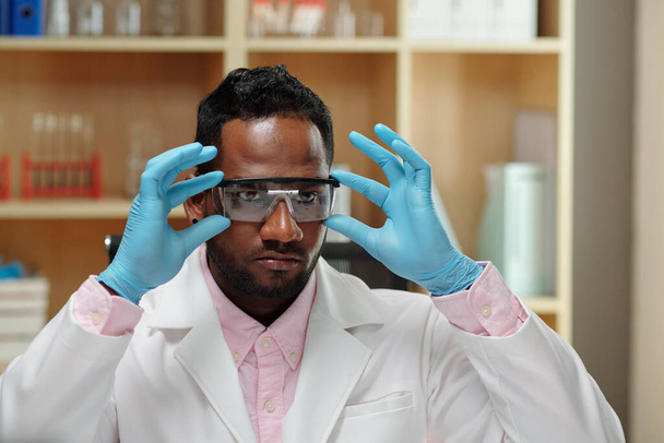 Joven hombre negro con bata de laboratorio y guantes que se pone anteojos protectores antes de llevar a cabo una investigación científica o trabajar con muestras tóxicas - Foto, Imagen
